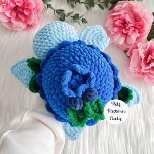 May include: A crocheted blueberry plush toy with blue, green, and light blue yarn. The toy has a round body with leaves and a stem. The text "Pdf Pattern Only" is on a white circle.