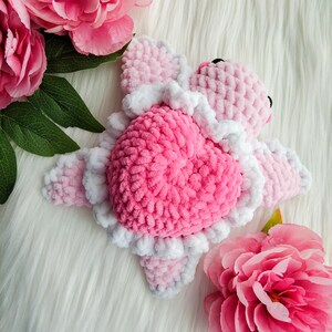 May include: Pink and white crocheted heart-shaped stuffed animal with a ruffled edge. The animal is lying on a white fluffy surface with pink flowers.