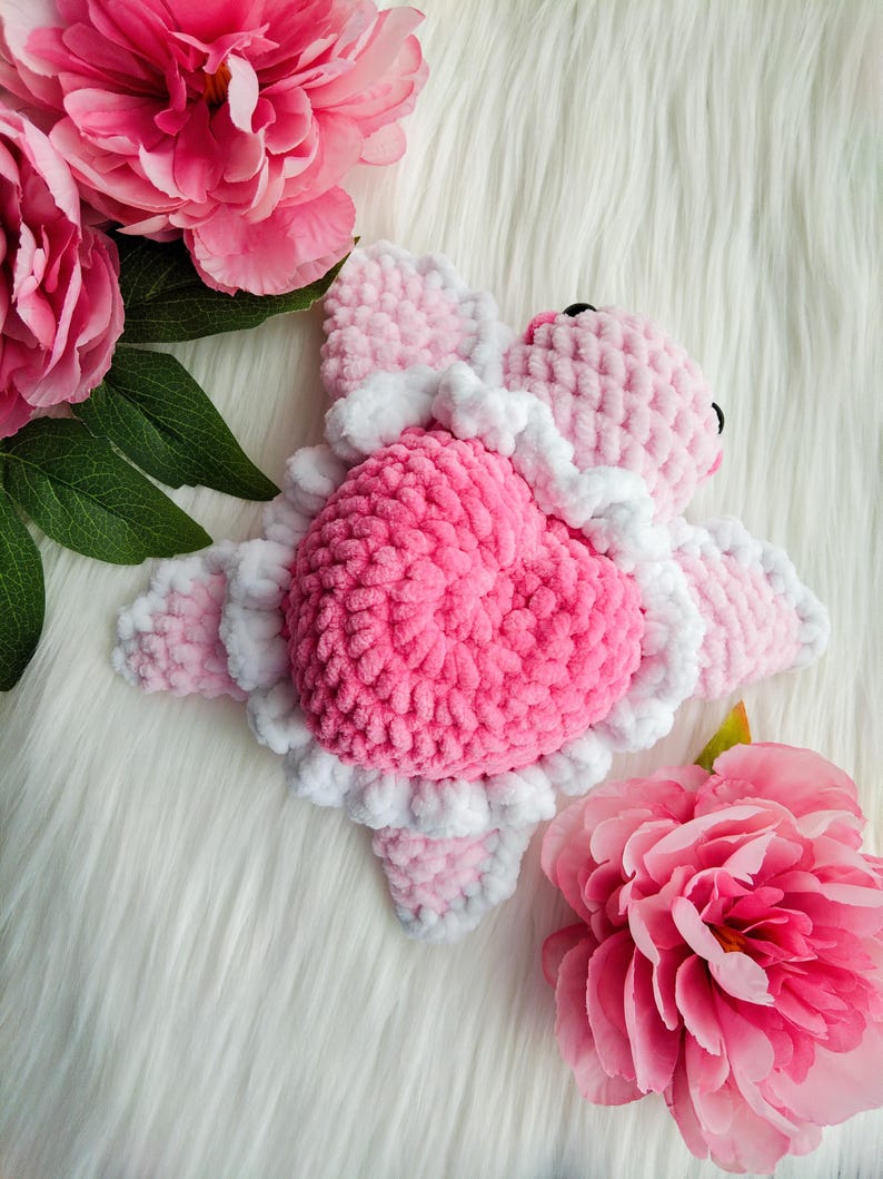 May include: Pink and white crocheted heart-shaped stuffed animal with a ruffled edge. The animal is lying on a white fluffy surface with pink flowers.
