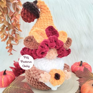 May include: A crocheted gnome figure featuring a tall orange hat topped with a dark brown acorn, adorned with layered burgundy and orange leaves. The gnome has a white beard, a peach-colored nose, and holds a small orange flower. Text reads Pdf Pattern Only.