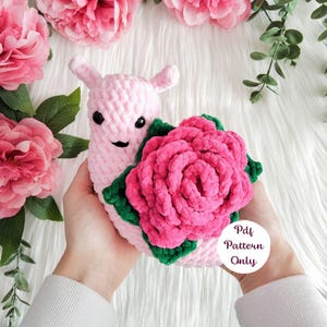 Könnte beinhalten: Ein handgefertigtes rosa Häkelschnecken-Spielzeug mit einer großen rosa Rose und grünen Blättern. Die Schnecke hat kleine schwarze Augen und einen lächelnden Mund. Der Text "Pdf Pattern Only" ist auf dem Bild. Es ist ein weiches, plüschiges Spielzeug.