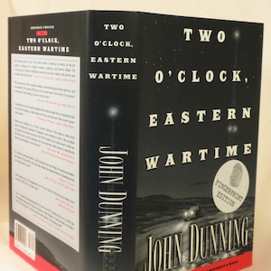 Puede incluir: Portada del libro "Two O'Clock, Eastern Wartime" de John Dunning. La portada presenta una imagen en blanco y negro de un coche conduciendo por una carretera de noche. El título del libro está en blanco sobre fondo negro. El nombre del autor está en blanco sobre fondo negro.