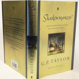 May include: A gold and black book cover with the title "Shadowmancer" and the author's name "G.P. Taylor" in a stylized font. The cover features a painting of a crumbling stone building with a dark sky in the background. The text "From darkness comes an ancient evil..." is printed below the title.