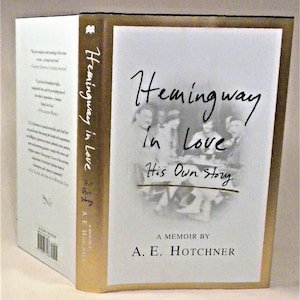 以下が含まれることがあります： 白い表紙に金色のカバーが付いた本。表紙には、アーネスト・ヘミングウェイとその友人たちの白黒写真が掲載されている。本のタイトルは「ヘミングウェイ・イン・ラブ：ヒズ・オウン・ストーリー」で、著者はA.E. ホットチナーである。