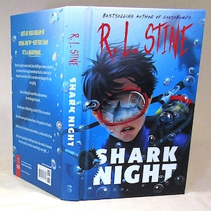 Puede incluir: Un libro de tapa dura titulado "Shark Night" de R.L. Stine. La portada presenta a una persona con una máscara de buceo con un tiburón visible en la lente. El fondo es azul con burbujas.