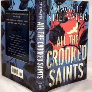 May include: A book cover with a blue background and a black rose. The title "All the Crooked Saints" is written in white letters. The author's name, "Maggie Stiefvater", is written in white letters above the title. The book is a #1 New York Times bestseller.