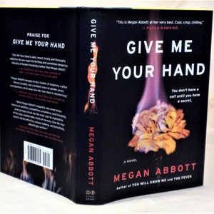 Może przedstawiać: Czarna okładka książki zatytułowana Give Me Your Hand autorstwa Megan Abbott. Z przodu znajduje się płonący żółty kwiat z fioletowymi płomieniami. Z tyłu znajdują się cytaty, kod kreskowy i logo Little, Brown. Grzbiet zawiera tytuł i autora.