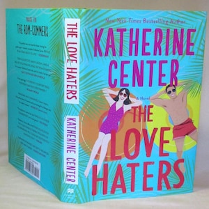 Könnte beinhalten: Ein Hardcover-Buch mit dem Titel "The Love Haters" von Katherine Center. Das Cover zeigt Illustrationen einer Frau in einem pinkfarbenen Badeanzug und eines Mannes in roten Shorts, beide mit Sonnenbrille, vor einem türkisfarbenen Hintergrund.