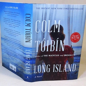 May include: Book cover for "Long Island" by Colm Tóibín. The cover features a woman looking out a window at the ocean. The title of the book is in large, white letters on a blue background. The author's name is in smaller white letters below the title. The book is a novel and is an Oprah's Book Club selection.