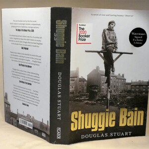 May include: A black and white photograph of a young boy sitting on a metal bar. The photo is on the cover of a book titled "Shuggie Bain" by Douglas Stuart. The book was shortlisted for the 2020 Booker Prize.