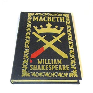 May include: A hardcover book titled "Macbeth" by William Shakespeare. The cover is black with gold lettering and a gold illustration of a crown and crossed swords. A red blood stain is on one of the swords.