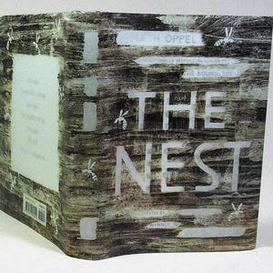 以下が含まれることがあります： 木目調のデザインが施された「The Nest」というタイトルのハードカバー本。表紙には大きな白い文字と蜂のイラストが描かれています。著者の名前やその他のテキストも表示されています。