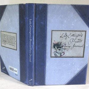 May include: A hardback book titled "Lady Cottington's Pressed Fairy Journal." The book features a light blue cover with dark blue detailing and a framed fairy illustration. The spine reads "Lady Cottington's Pressed Fairy Journal."