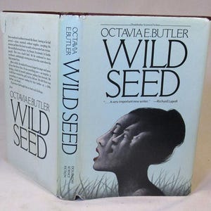 Puede incluir: Un libro de tapa dura titulado "WILD SEED" de Octavia E. Butler. La portada presenta una ilustración en blanco y negro del rostro de una mujer con múltiples perfiles superpuestos. El título y el nombre del autor aparecen en la portada y el lomo.