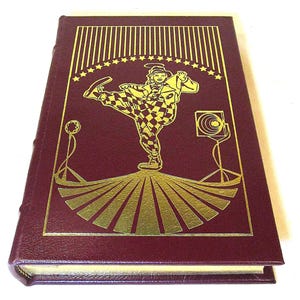 May include: A burgundy book with a gold illustration of a clown in a checkered outfit, kicking up his leg. The cover also features a spotlight and a star-studded banner. The book has gold page edges.