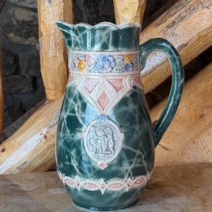 May include: A dark green ceramic pitcher featuring a marbled finish, a scalloped rim, and a curved handle. The body is decorated with a central white circular relief depicting figures, accented by intricate geometric patterns and floral motifs.