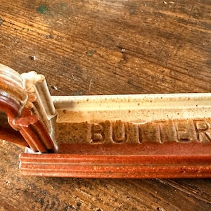May include: A ceramic butter dish with a rustic design. The dish is rectangular with a curved handle and the word "BUTTER" embossed on the front. The colors are shades of brown, cream, and red.