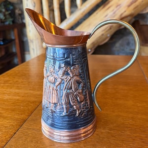 May include: A copper and brass pitcher featuring a relief design of two figures in traditional attire. The vessel has a polished copper rim and base, a dark oxidized body with embossed detailing, and a curved brass handle attached to the side.