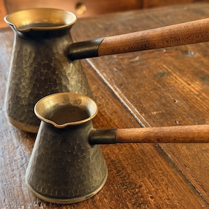 May include: Two copper coffee pots with wooden handles. The pots have a hammered texture and a brass-colored rim. The wooden handles are attached with a metal band and have a small metal ring at the end.