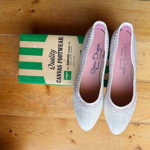 May include: A pair of white canvas flats with a multicolored woven trim around the opening and pink insoles labeled Spice Drops. The shoes are positioned next to a green and white striped box labeled Quality Canvas Footwear by The Servus Rubber Co.