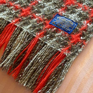 May include: A woven wool blanket featuring a brown, white, and bright orange plaid pattern with long fringe edges. A blue rectangular label is stitched onto the fabric, reading Pendleton, Warranted to be a Pendleton, Pure Virgin Wool, Made in U.S.A.