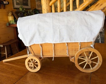 May include: A wooden model of a covered wagon featuring a light-colored natural wood frame, four spoked wheels, and a white fabric canopy secured with white string ties. The wagon includes a long wooden tongue extending from the front.