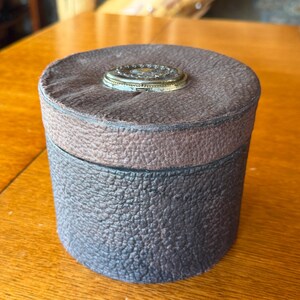 May include: A cylindrical dark brown leather box with a textured surface and a removable lid. The top of the lid features a centered, circular metallic ornament with a decorative embossed pattern and a smooth, light-colored center disc.