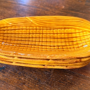 May include: A stack of ceramic corn-shaped serving dishes. The dishes are a golden yellow color with brown detailing, mimicking the appearance of corn on the cob. The dishes are stacked on top of each other.