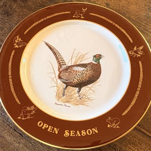 May include: A decorative plate with a brown rim and gold trim. The center features a detailed illustration of a pheasant in a grassy field. The words "OPEN SEASON" are printed in gold.