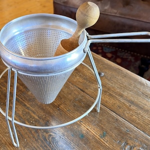 May include: A vintage metal food mill with a perforated cone-shaped strainer and a wooden pestle. The mill has a handle and is supported by a wire frame. The wooden pestle is light brown. The food mill is designed for straining and pureeing food.