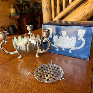 May include: A silver-toned metal lotus flower candle holder with two candle cups on curved arms, accompanied by a circular metal flower frog with perforated holes and a blue box labeled with the text "trompe l'oeil silver".