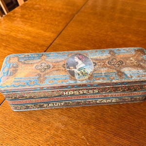 May include: Rectangular vintage Hostess Fruit Cake tin with a decorative design in blue, gold, and red. The tin features an illustration of a woman and the words "HOSTESS FRUIT CAKE".