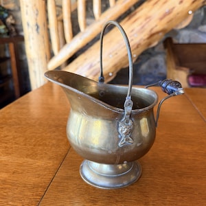 May include: A vintage brass coal scuttle with a handle and a small spout. The scuttle has a decorative fish emblem on the front and a black handle on the side. The piece is sitting on a wooden table.