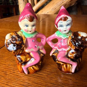 May include: A pair of vintage ceramic elf figurines, each with a pink outfit, green collar, and a red hat. The elves are perched on a brown tree stump. The figurines have detailed facial features and are likely salt and pepper shakers.