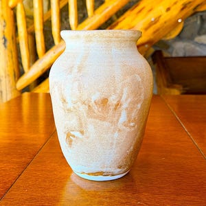 May include: A beige ceramic vase with a wide body and a narrow neck. The vase has a mottled, textured surface with brown and white tones. It sits on a polished wooden table, showcasing its rustic charm.