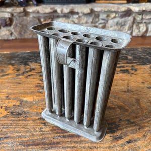 May include: A vintage metal candle mold featuring a rectangular frame with twelve cylindrical tubes arranged in two rows. The gray, weathered tin structure includes a flat base, a top plate with circular openings, and a small metal handle on the side.