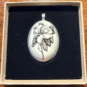 May include: An oval pendant with a detailed black and white floral design, likely a magnolia, set against a cream-colored background. The pendant is in a silver-colored setting and is presented in a small, open gift box with a black interior.