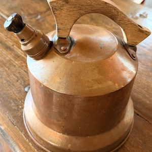 May include: A copper tea kettle with a wooden handle and spout. The kettle has a round body and a wooden handle. The spout has a small, dark knob. The kettle is made of copper and has a shiny finish.