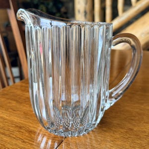 May include: A clear glass pitcher with vertical ribbed detailing. The pitcher has a curved handle and a spout for easy pouring. The pitcher is sitting on a wooden surface. The pitcher is likely used for serving beverages.