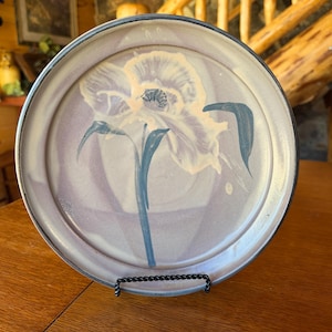 May include: A round ceramic plate featuring a hand-painted floral design in shades of cream, soft purple, and deep blue-green. The plate has a smooth, glazed finish with a dark, defined rim and a central iris-like flower motif with long stems.