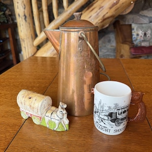 May include: A tall copper coffee percolator with a lid and handle, a ceramic covered wagon figurine with a horse, and a white ceramic mug featuring a black graphic of a stagecoach and the text Straight Shooter with a brown pistol-shaped handle.