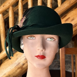 May include: A dark green cloche hat with a rounded crown and a downturned brim. The hat is adorned with a feather and ribbon detail. The hat is displayed on a mannequin head.
