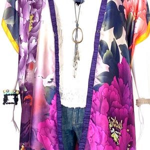 May include: A long, purple and pink floral print kimono with a white background. The kimono has a purple trim and is open in the front. The kimono is worn over a white shirt and blue jeans.