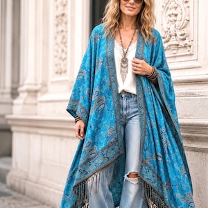 May include: A bright blue open-front kimono featuring a detailed dark blue and brown paisley pattern, wide sleeves, and a long fringe hem. The garment is worn over a white camisole with lace trim and light-wash distressed denim jeans.