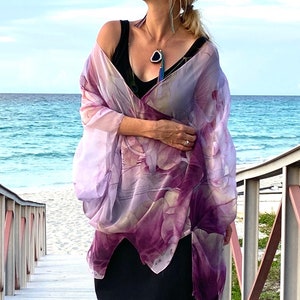 May include: A woman wearing a purple and white floral print silk scarf with a black tank top underneath. The scarf is draped over her shoulders and tied at the front. She is standing on a wooden walkway with a beach in the background.
