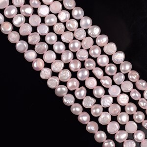 Puede incluir: Primer plano de varios hilos de perlas de agua dulce de color rosa claro. Las perlas tienen forma irregular y un brillo suave. El fondo es negro, lo que hace que las perlas destaquen.