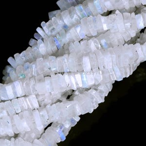 Puede incluir: Un hilo de cuentas de piedra lunar blanca translúcida con forma de pequeños rectángulos planos. Cada cuenta muestra un sutil brillo azul iridiscente que capta la luz, creando un efecto brillante a lo largo de toda la longitud del hilo.