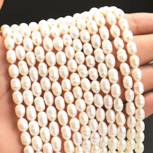 Puede incluir: Un hilo de perlas de agua dulce blancas de forma ovalada, dispuestas en varias filas paralelas. Cada perla presenta una superficie lisa y brillante con sutiles variaciones en forma y tamaño, reflejando la luz en toda la colección de cuentas.