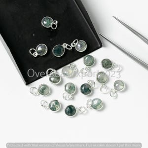Puede incluir: Una colección de colgantes redondos plateados con un centro de piedra verde y transparente. Los colgantes están esparcidos sobre una superficie blanca con una bandeja negra y unas pinzas plateadas. El texto "Oversea Export 2023" es visible.