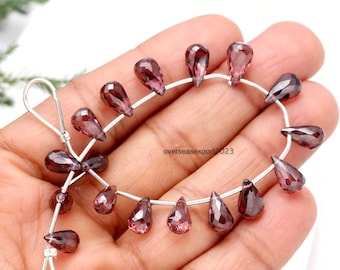 May include: A string of faceted, teardrop-shaped, dark red-purple gemstones strung on silver wire. The translucent beads have a polished finish. The gemstones are displayed against a light background.